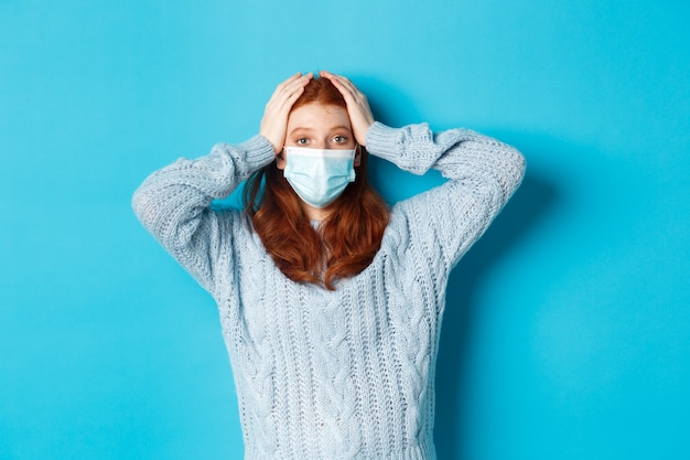 Invierno, covid-19 y concepto de distanciamiento social. Chica pelirroja preocupada en mascarilla mirando angustiado, tomados de la mano en la cabeza en pánico, de pie sobre fondo azul.