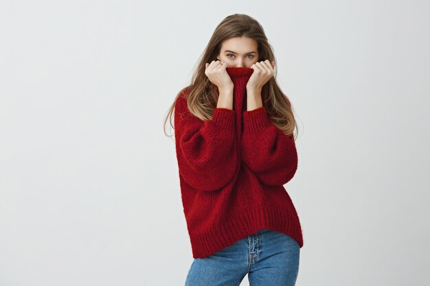 El invierno está cerca. Mujer delgada y atractiva con un suéter holgado de moda que oculta la cara en el cuello mientras mira, siente frío o se sonroja de cumplidos, de pie.