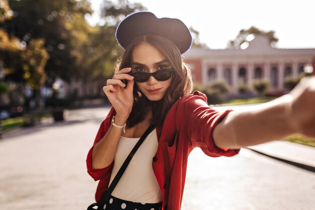 Involucrar a la joven parisina con una elegante boina de cabello ondulado oscuro y gafas de sol en la parte superior blanca y una camisa roja haciendo selfie contra el fondo de la ciudad Clima soleado y cálido