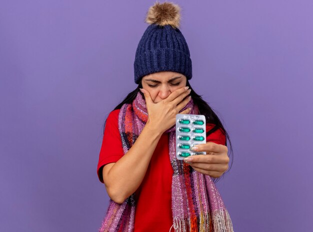 Foto gratuita irritada joven caucásica enferma con gorro de invierno y bufanda estirando paquete de cápsulas hacia la cámara manteniendo la mano en la boca con los ojos cerrados aislado sobre fondo púrpura con espacio de copia