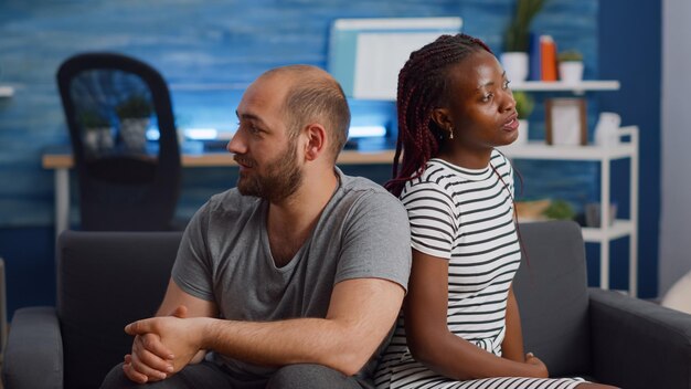 Irritada pareja interracial peleando y sentado en el sofá en casa. Amantes multiétnicos con argumento gritando en la sala de estar. Gente de raza mixta enojada con problemas de relación gritando