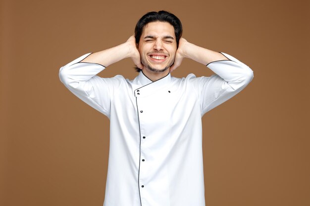 Irritado joven chef masculino vistiendo uniforme cerrando los oídos con las manos con los ojos cerrados aislado sobre fondo marrón.