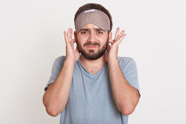 Irritado joven enojado con barba, mirando directamente cubriendo sus orejas con los dedos, vistiendo camiseta y máscara para dormir