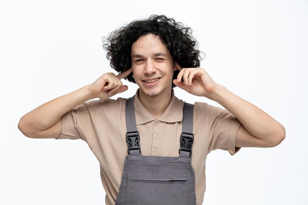 Irritado joven trabajador de la construcción vistiendo uniforme mirando a la cámara cerrando los oídos con los dedos aislado sobre fondo blanco.