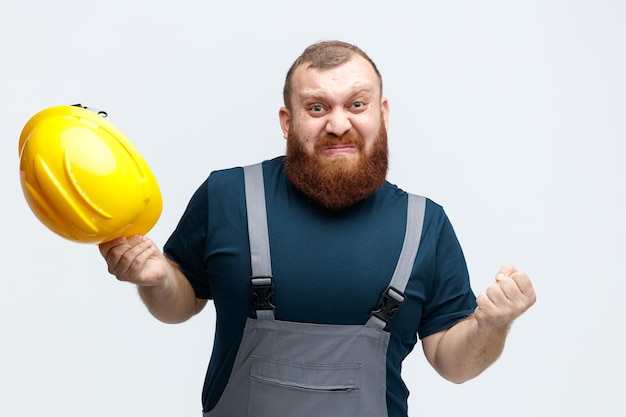 Irritado joven trabajador de la construcción vistiendo uniforme sosteniendo casco de seguridad manteniendo el puño en el aire mirando a la cámara aislada sobre fondo blanco.