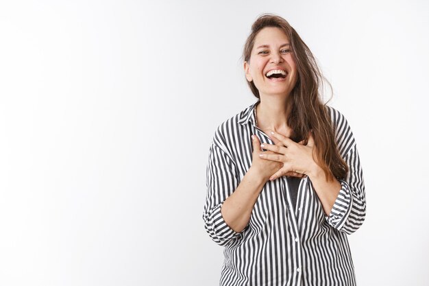 Ja, ja, tan divertido. Retrato de encantadora mujer de mediana edad despreocupada en blusa a rayas tomados de la mano en el pecho tocado y complacido riendo en voz alta asombrado y entretenido de pie sobre la pared blanca