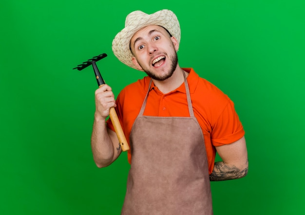 Foto gratuita jardinero alegre hombre vestido con sombrero de jardinería tiene rastrillo y sostiene la mano detrás de la espalda