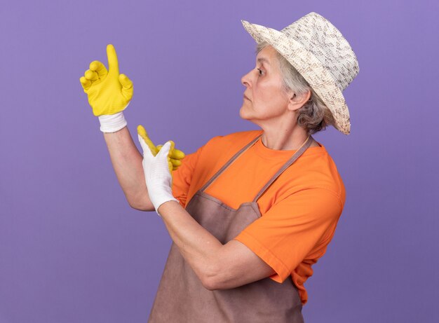 Jardinero anciano ansioso vistiendo gorro y guantes de jardinería apuntando hacia arriba mirando al lado en púrpura