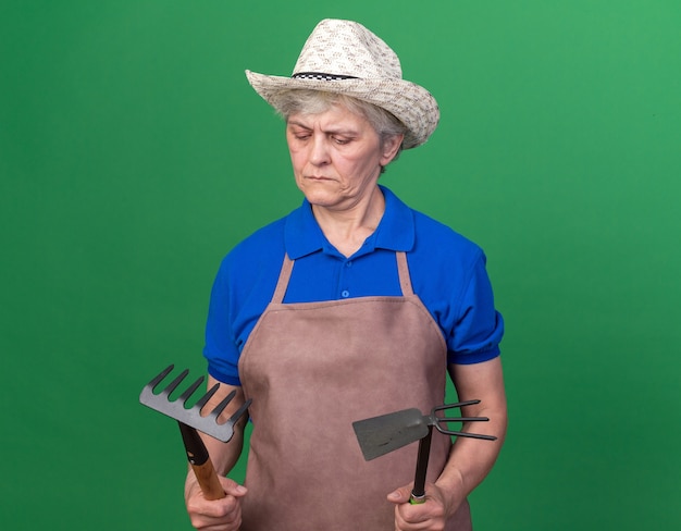 Foto gratuita jardinero anciano confundido con sombrero de jardinería sosteniendo rastrillo y rastrillo azada en verde