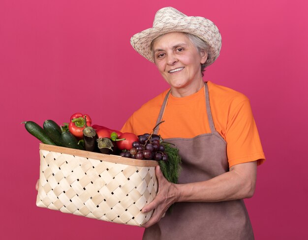 Jardinero anciano sonriente vistiendo sombrero de jardinería sosteniendo canasta de verduras aislado en la pared rosa con espacio de copia