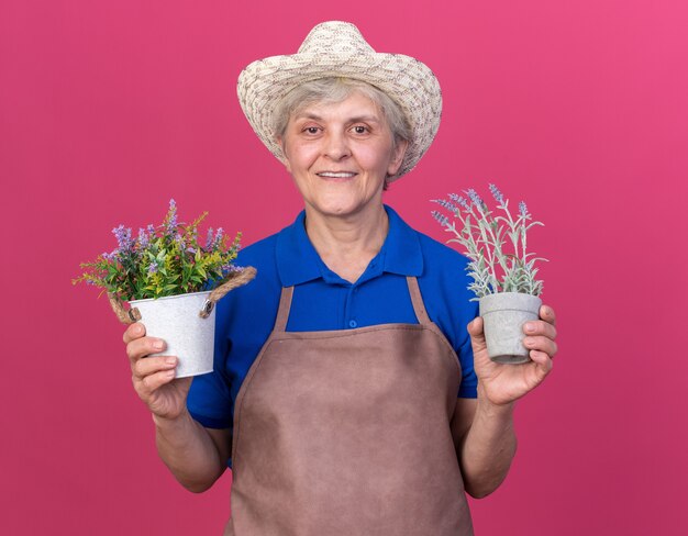 Jardinero anciano sonriente vistiendo sombrero de jardinería sosteniendo macetas