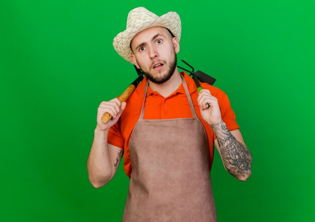 Jardinero despistado hombre vestido con sombrero de jardinería tiene herramientas de jardinería sobre los hombros