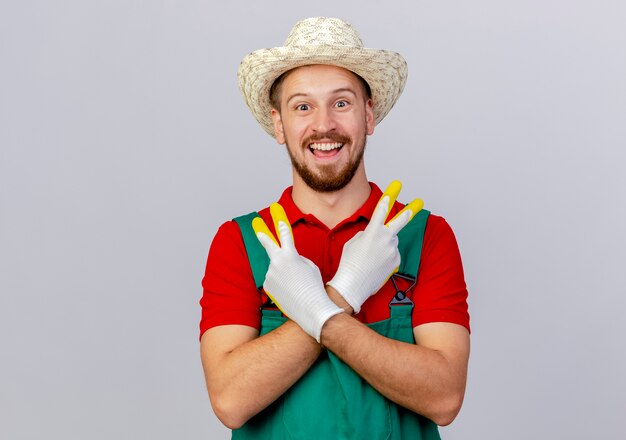 Jardinero eslavo guapo joven alegre en uniforme con guantes de jardinería y sombrero mirando manteniendo las manos cruzadas haciendo el signo de la paz aislado