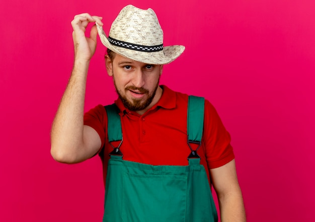 Jardinero eslavo guapo joven confiado en uniforme y sombrero mirando agarrando sombrero