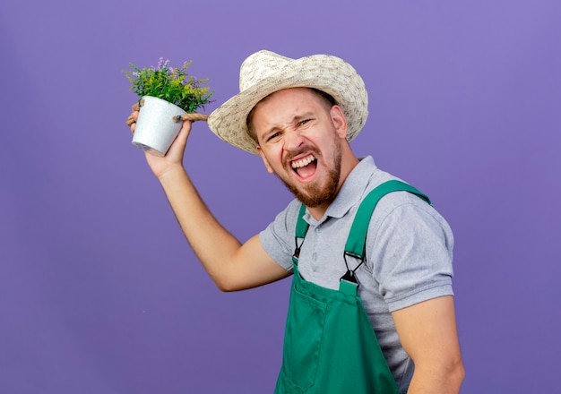 Jardinero eslavo guapo joven confiado en uniforme y sombrero de pie en la vista de perfil sosteniendo maceta mirando aislado en la pared púrpura con espacio de copia