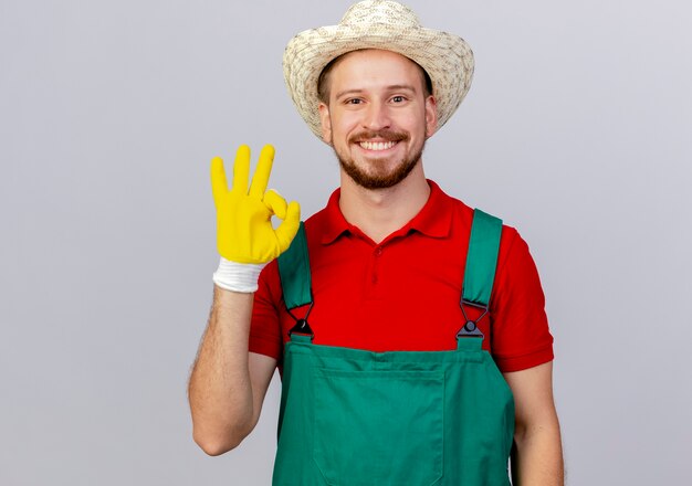 Jardinero eslavo guapo joven feliz en uniforme con guantes de jardinería y sombrero haciendo bien signo aislado en la pared blanca con espacio de copia