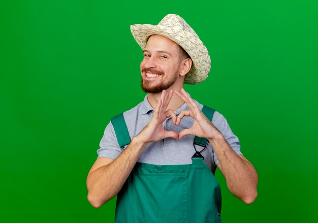 Jardinero eslavo guapo joven feliz en uniforme y sombrero mirando haciendo signo de corazón