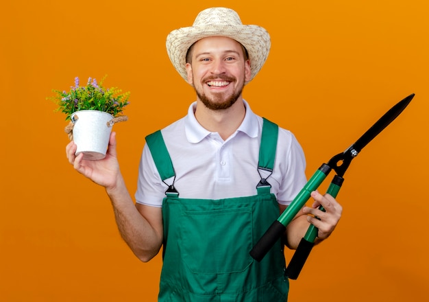 Jardinero eslavo guapo joven feliz en uniforme y sombrero con planta de flores y podadoras mirando aislado