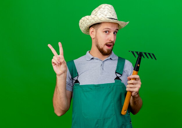 Jardinero eslavo guapo joven impresionado en uniforme y sombrero mirando sosteniendo rastrillo y haciendo el signo de la paz