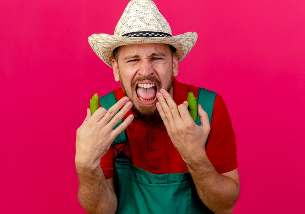 Jardinero eslavo guapo joven irritado en uniforme y sombrero sosteniendo mitades de pimienta mirando