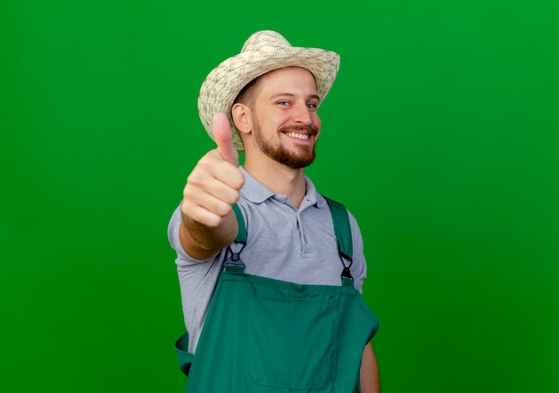 Foto gratuita jardinero eslavo guapo joven sonriente en uniforme y sombrero mirando mostrando el pulgar hacia arriba