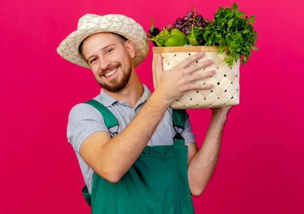 Jardinero eslavo guapo joven sonriente en uniforme y sombrero sosteniendo la cesta de verduras cerca de la cabeza mirando