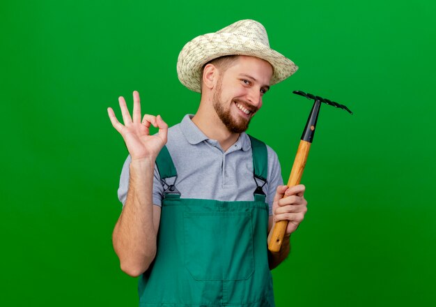 Jardinero eslavo guapo joven sonriente en uniforme y sombrero sosteniendo rastrillo mirando haciendo bien signo aislado en la pared verde con espacio de copia