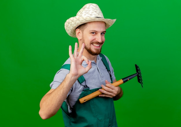 Jardinero eslavo guapo joven sonriente en uniforme y sombrero sosteniendo rastrillo mirando haciendo bien signo aislado en la pared verde