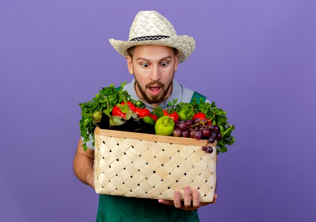 Jardinero eslavo hermoso joven sorprendido en uniforme y sombrero que sostiene y que mira la cesta de verduras aislada