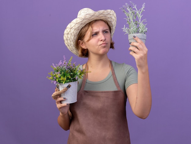 Jardinero eslavo joven pensativo con sombrero de jardinería sosteniendo y mirando flores en macetas en púrpura