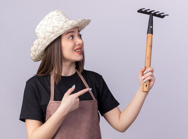 Foto gratuita jardinero femenino bastante caucásico ansioso con sombrero de jardinería sosteniendo y apuntando al rastrillo aislado en la pared blanca con espacio de copia