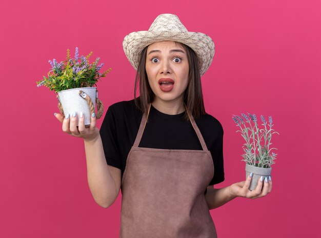 Jardinero femenino bastante caucásico ansioso vistiendo sombrero de jardinería sosteniendo macetas en rosa