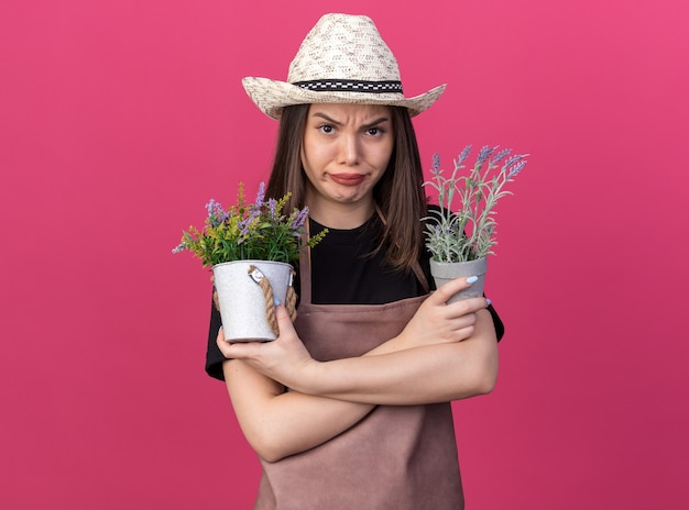 Jardinero femenino bastante caucásico enojado con sombrero de jardinería se encuentra con los brazos cruzados sosteniendo macetas