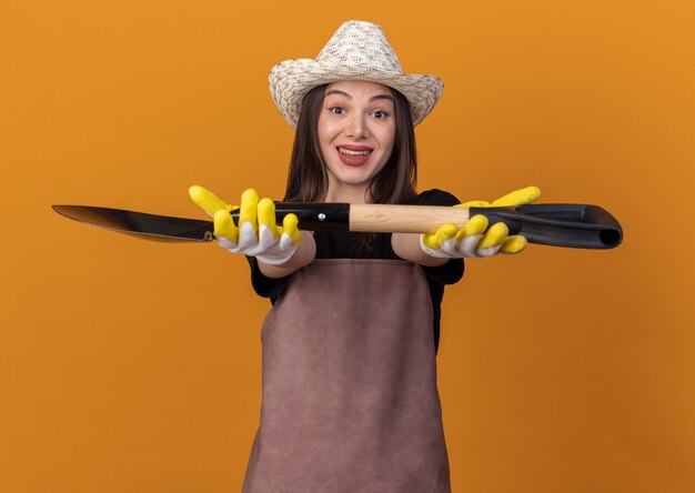 Jardinero femenino bastante caucásico impresionado con sombrero de jardinería y guantes sosteniendo la pala en naranja