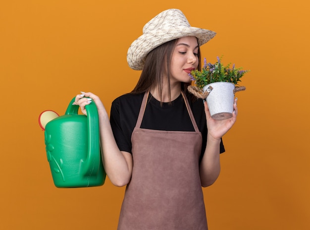 Jardinero femenino bastante caucásico satisfecho con sombrero de jardinería sosteniendo regadera y oliendo flores en maceta aislado en pared naranja con espacio de copia