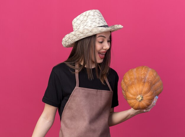 Jardinero femenino bastante caucásico sorprendido que lleva el sombrero que cultiva un huerto que sostiene y que mira la calabaza en rosa