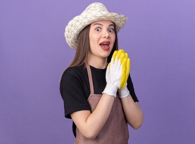 Jardinero femenino bastante caucásico sorprendido con sombrero de jardinería y guantes tomados de la mano juntos en púrpura