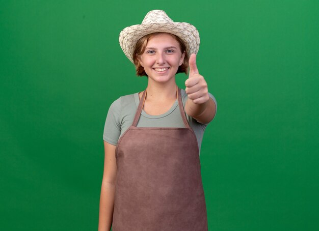 Jardinero femenino eslavo joven sonriente que lleva el sombrero que cultiva un huerto pulgar hacia arriba en verde