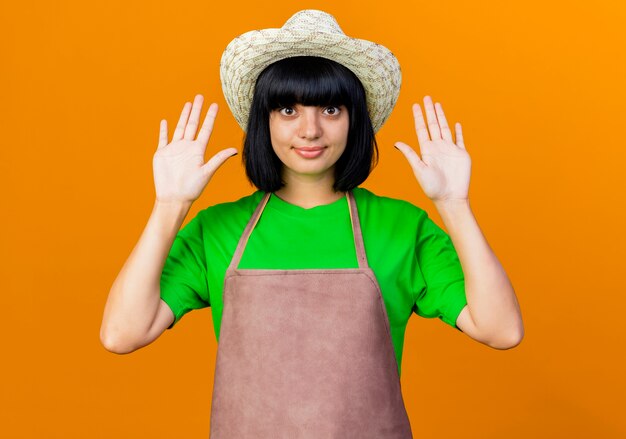 Jardinero femenino joven complacido en uniforme vistiendo sombrero de jardinería se encuentra con las manos levantadas