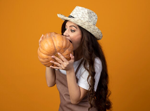 Jardinero femenino joven impresionado en uniforme con sombrero de jardinería sostiene y pretende morder calabaza aislado en la pared naranja con espacio de copia