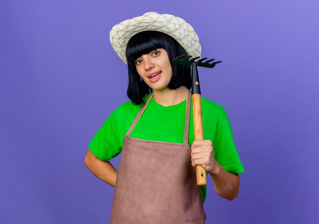 Foto gratuita jardinero joven alegre en uniforme vistiendo sombrero de jardinería tiene rastrillo