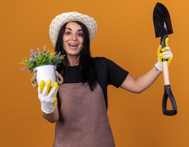 Jardinero joven alegre vestida con uniforme y sombrero con guantes de jardinero sosteniendo la pala y la maceta mirando al frente aislado en la pared naranja