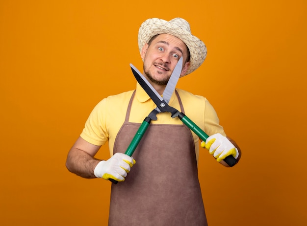 Jardinero joven divertido vistiendo mono y sombrero sosteniendo cortasetos mirando al frente sonriendo con cara feliz de pie sobre la pared naranja