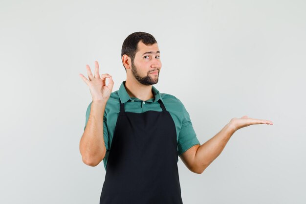 Jardinero joven que muestra un gesto aceptable mientras levanta la palma de su mano en camiseta, delantal y parece complacido. vista frontal.