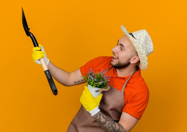 Jardinero macho alegre vistiendo guantes y gorro de jardinería sostiene flores en maceta y pala mirando al lado