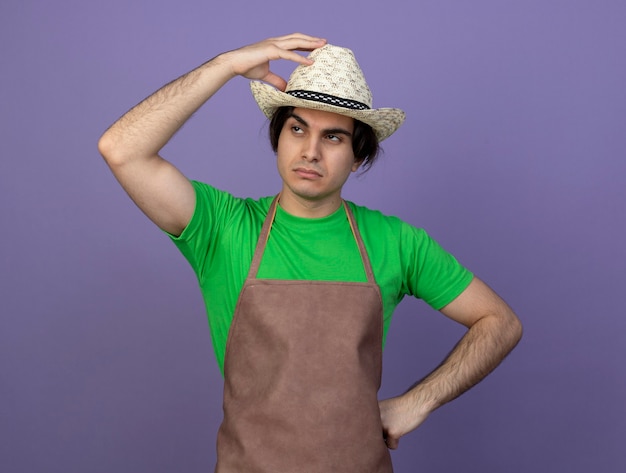 Jardinero macho joven cansado en uniforme vistiendo sombrero de jardinería poniendo la mano en el sombrero