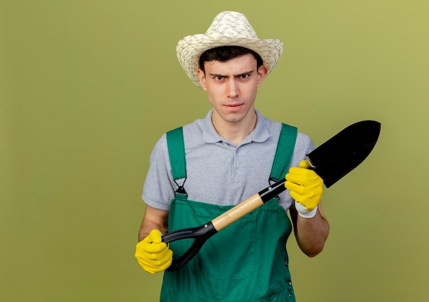 Jardinero macho joven disgustado vistiendo guantes y gorro de jardinería tiene pala