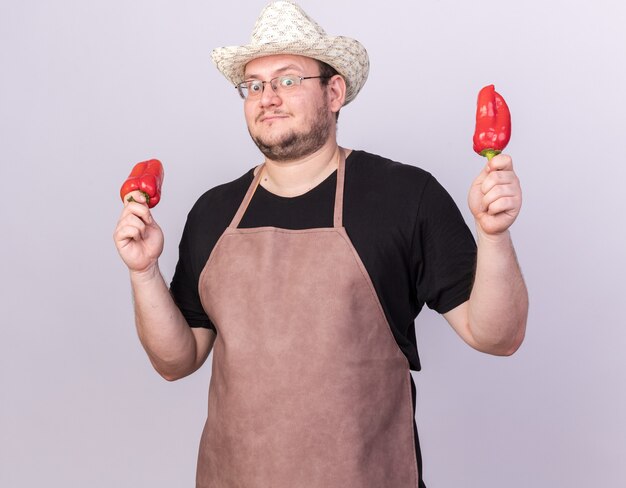 Jardinero macho joven impresionado con sombrero de jardinería sosteniendo pimientos aislados en la pared blanca