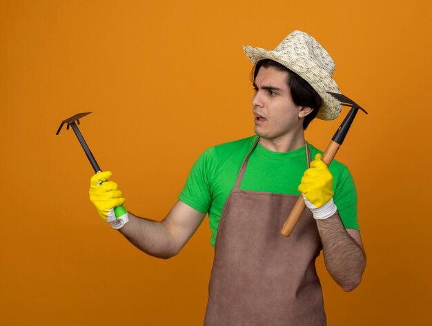 Jardinero macho joven pensativo en uniforme con sombrero de jardinería con guantes sosteniendo y mirando azada rastrillo con rastrillo aislado en naranja