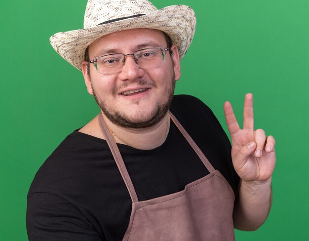 Jardinero macho joven sonriente con sombrero de jardinería mostrando gesto de paz aislado en la pared verde